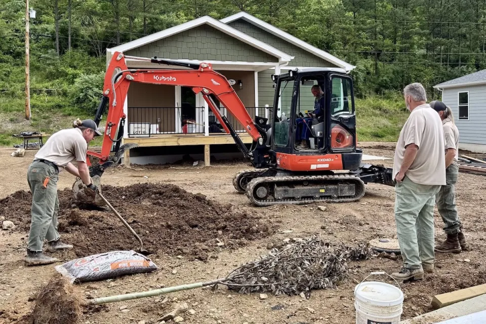 Small green house on unfinished lot with red earthmover and workers in front yard.
